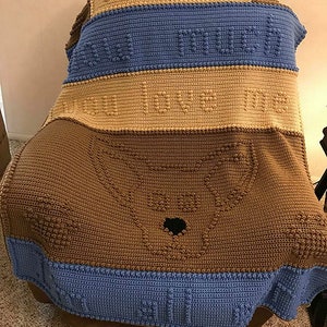 May include: A handmade crochet blanket draped over a chair. The blanket features a brown dog face design and the words "I love you so much" in a textured stitch. The blanket has blue and tan stripes.