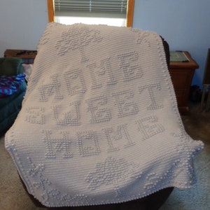 May include: A white crocheted blanket draped over a chair. The blanket features the words "HOME SWEET HOME" in raised letters, along with tree and star designs. The blanket is made of a soft, textured material.