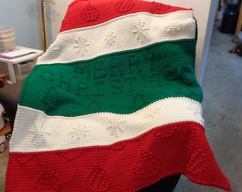 May include: A crocheted blanket with a red, white, and green striped pattern. The blanket features a Christmas theme with snowflakes, ornaments, and the words "Merry Christmas".