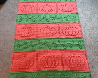 May include: A crocheted blanket with a repeating pattern of pumpkins and leaves in shades of green and orange.