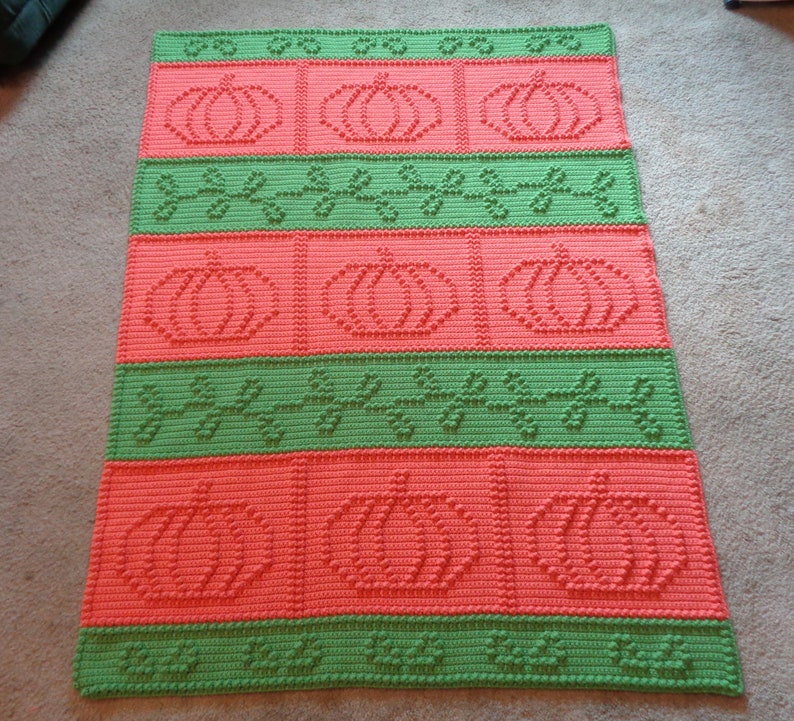 May include: A crocheted blanket with a repeating pattern of pumpkins and leaves in shades of green and orange.