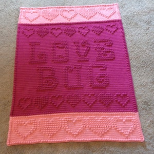 May include: A pink and red crocheted blanket with the word "LOVE BIG" in raised letters. The blanket has a border of pink hearts.