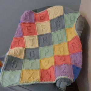 Puede incluir: Una manta de bebé de crochet colorida con cada cuadrado que presenta una letra diferente del alfabeto. La manta está hecha en tonos de rosa, amarillo, azul y verde.