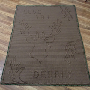 Op de afbeelding: Een bruin gebreid deken met een groene rand. Het deken heeft een hert ontwerp met de woorden "LOVE YOU" en "DEERLY" in het midden.
