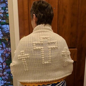 May include: White crocheted shawl with a cross pattern and a yellow crocheted border. The shawl is worn by a person.