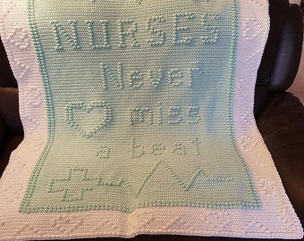 Peut inclure: Une couverture au crochet faite à la main, blanche et vert menthe. La couverture présente les mots "Nurses Never Miss a Beat" en lettres en relief, ainsi qu'un cœur et un symbole de croix médicale. La bordure a un motif texturé.