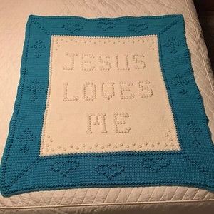 Pode incluir: Um cobertor de bebé branco de crochê com uma borda azul. O cobertor tem as palavras "Jesus ama-te" em fio branco.