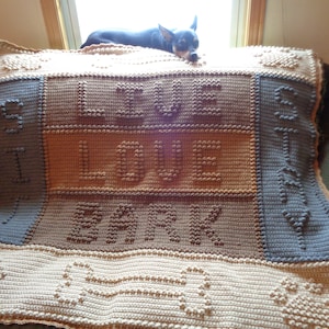 May include: A crocheted blanket with the words "I love dogs" in brown and tan yarn. The blanket is made with a textured stitch and has a border of bone-shaped designs. A small black dog is lying on the blanket.