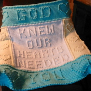 May include: A crocheted blanket in shades of white, light blue, and cream. The blanket features the words "GOD KNEW OUR HEARTS NEEDED YOU" in raised lettering. Heart-shaped accents are also incorporated into the design.