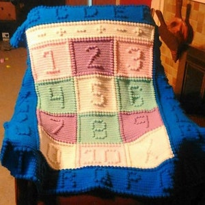 Può includere: Una copertina per neonati in crochet blu e bianca con un design colorato di alfabeto e numeri. La copertina ha un bordo blu con pompon bianchi.