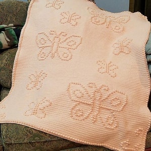May include: A peach-colored crocheted blanket with a raised butterfly design. The blanket is made with a soft, textured yarn and features a repeating pattern of butterflies.