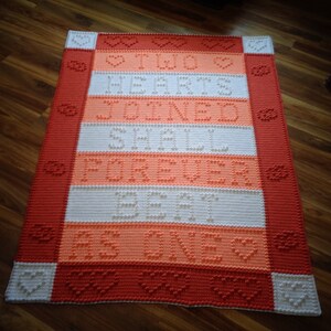 Könnte beinhalten: Eine gehäkelte Decke in Rot, Weiß und Orange. Die Decke hat ein sich wiederholendes Muster aus Herzen und den Worten "Two Hearts Joined Forever Be One".