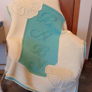 May include: A crocheted baby blanket with a white and light blue design. The blanket features a scalloped edge and a textured pattern.