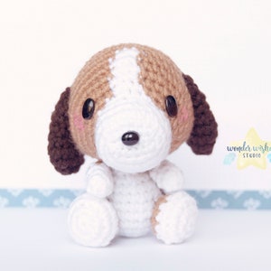 Pode incluir: Um pequeno brinquedo de c&atilde;o de croch&ecirc; com pelo marrom e branco. O c&atilde;o tem uma cara branca com olhos marrons e um nariz preto.