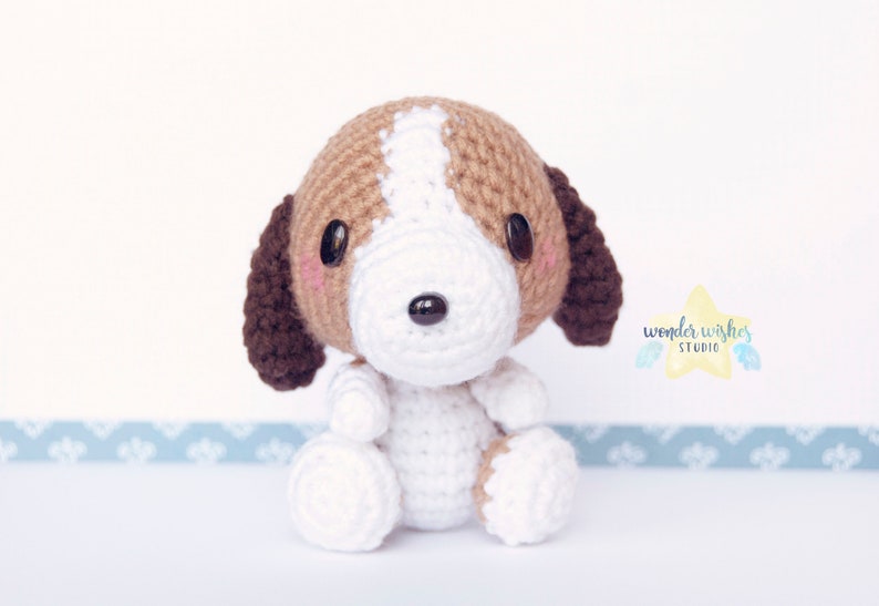 Pode incluir: Um pequeno brinquedo de c&atilde;o de croch&ecirc; com pelo marrom e branco. O c&atilde;o tem uma cara branca com olhos marrons e um nariz preto.