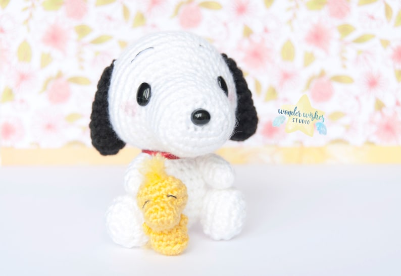 Pode incluir: Um c&atilde;o de pel&uacute;cia branco de croch&ecirc; com orelhas pretas, segurando um p&aacute;ssaro de pel&uacute;cia amarelo de croch&ecirc;. O c&atilde;o tem olhos pretos e uma coleira vermelha.
