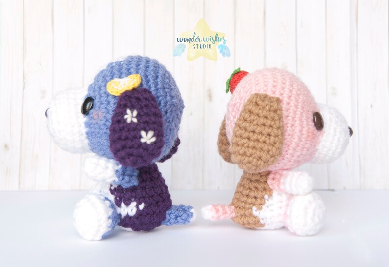Pode incluir: Dois c&atilde;es de pel&uacute;cia de croch&ecirc;, um azul e roxo com uma flor amarela na cabe&ccedil;a, e o outro rosa e marrom com um morango na cabe&ccedil;a.