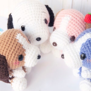 Pode incluir: Quatro brinquedos de pel&uacute;cia de cachorro de croch&ecirc; em diferentes cores e padr&otilde;es. Os c&atilde;es est&atilde;o sentados em c&iacute;rculo sobre uma superf&iacute;cie branca. Os c&atilde;es usam todos acess&oacute;rios diferentes, como um morango, um lim&atilde;o e uma flor.
