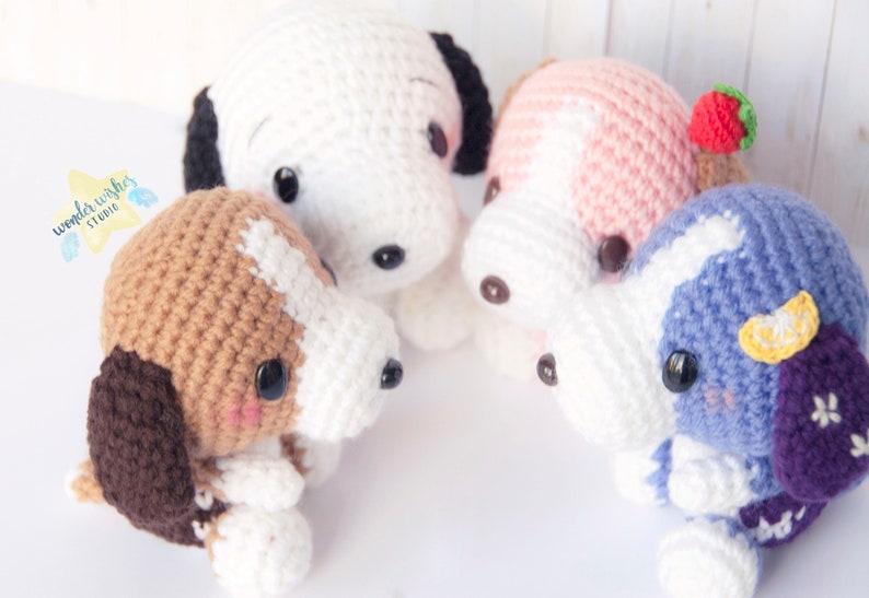 Pode incluir: Quatro brinquedos de pel&uacute;cia de cachorro de croch&ecirc; em diferentes cores e padr&otilde;es. Os c&atilde;es est&atilde;o sentados em c&iacute;rculo sobre uma superf&iacute;cie branca. Os c&atilde;es usam todos acess&oacute;rios diferentes, como um morango, um lim&atilde;o e uma flor.