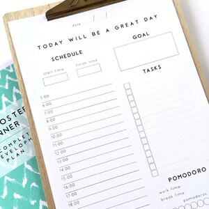 May include: A white printable daily schedule planner with a wooden clipboard. The planner includes sections for a goal, tasks, and a Pomodoro timer. The text "TODAY WILL BE A GREAT DAY" is at the top of the planner.