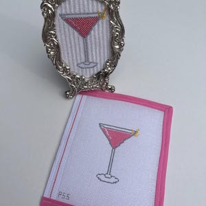 May include: A cross-stitch pattern of a pink cocktail in a glass with a yellow garnish, displayed in an ornate silver frame and on a white fabric square with pink trim, featuring the text P55 and a small red cross symbol in the corner.