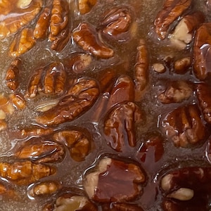 May include: A collection of whole brown pecan halves submerged in a thick, translucent amber-colored syrup. The nuts have a textured, ridged surface and are coated in a glossy, viscous liquid that fills the gaps between each individual nut.