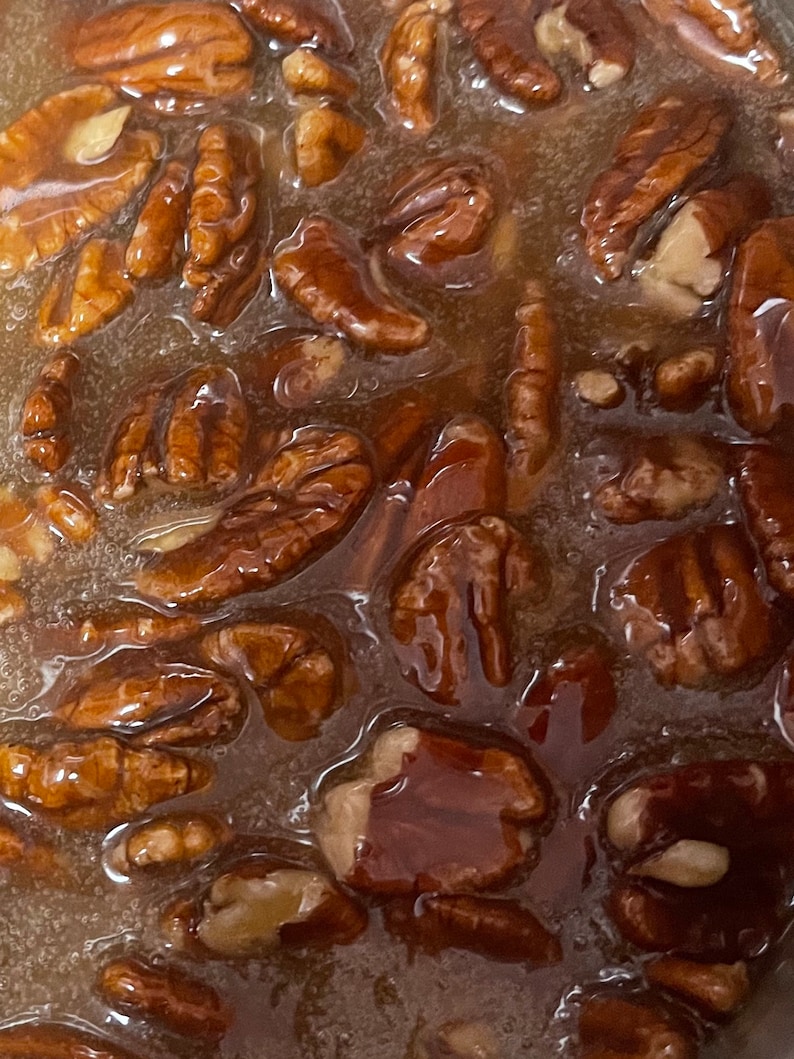 May include: A collection of whole brown pecan halves submerged in a thick, translucent amber-colored syrup. The nuts have a textured, ridged surface and are coated in a glossy, viscous liquid that fills the gaps between each individual nut.