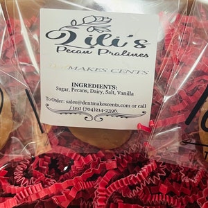 May include: Three individually wrapped pecan pralines in clear plastic bags with black text reading MADE with love THANK YOU. A central white label displays Lili's Pecan Pralines, DENT MAKES CENTS, ingredients, and contact information, all nestled in red shredded paper.