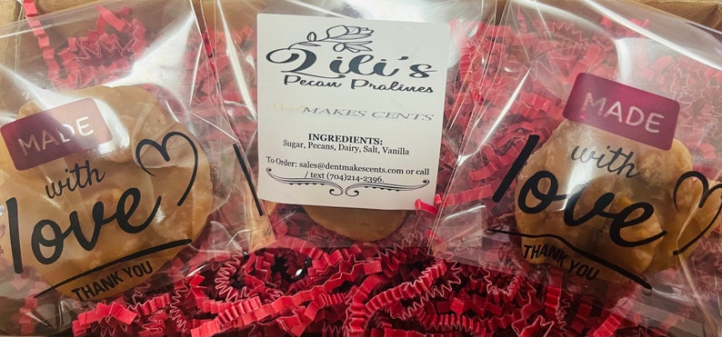 May include: Three individually wrapped pecan pralines in clear plastic bags with black text reading MADE with love THANK YOU. A central white label displays Lili's Pecan Pralines, DENT MAKES CENTS, ingredients, and contact information, all nestled in red shredded paper.
