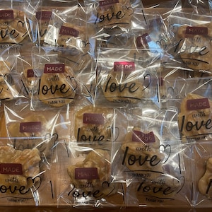May include: A collection of individually wrapped tan-colored nut brittle pieces in clear plastic bags. Each bag features a maroon rectangular label with the white text MADE with love and the black script text love, plus the words THANK YOU.