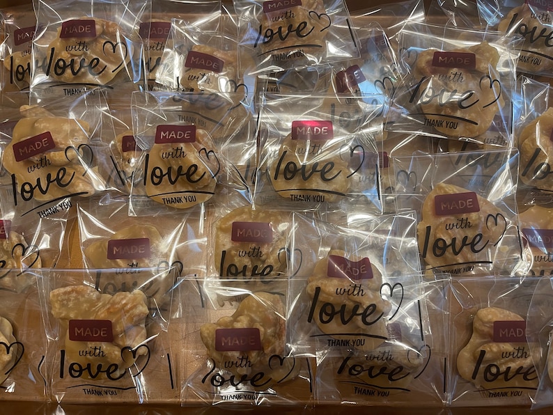 May include: A collection of individually wrapped tan-colored nut brittle pieces in clear plastic bags. Each bag features a maroon rectangular label with the white text MADE with love and the black script text love, plus the words THANK YOU.