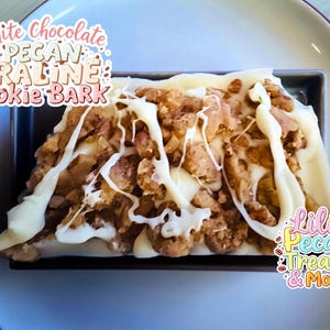 May include: White chocolate pecan praline cookie bark. The dessert features a rectangular shape with a base of cookie bark, topped with pecan pieces and drizzled with white chocolate. The image includes text that reads "White Chocolate Pecan Praline Cookie Bark."