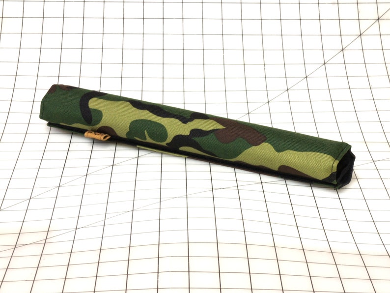 Puede incluir: Un tubo de tela con estampado de camuflaje, una costura negra y una etiqueta de cuero marr&oacute;n.