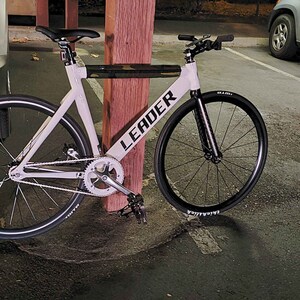 Puede incluir: Una bicicleta de pi&ntilde;&oacute;n fijo blanca con neum&aacute;ticos negros y un logotipo LEADER negro en el cuadro. La bicicleta est&aacute; apoyada en un poste de madera.
