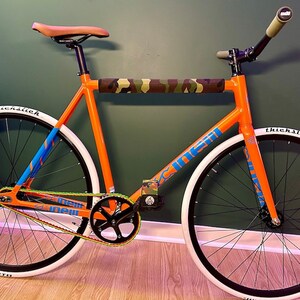 Puede incluir: Una bicicleta de pi&ntilde;&oacute;n fijo naranja y azul con neum&aacute;ticos blancos y una almohadilla de manillar de camuflaje. La bicicleta tiene un sill&iacute;n negro y una cadena negra.