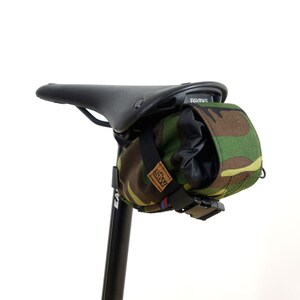 Può includere: Una borsa per sella da bicicletta in tessuto mimetico con cinturino e fibbia neri. La borsa è fissata a un reggisella nero con una sella Brooks nera.