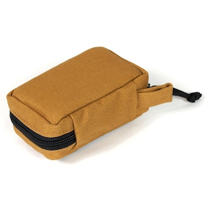 May include: A rectangular, tan-colored pouch with a black zipper closure. The pouch has a small fabric loop on one end and a black cord with a knot attached. The pouch is made of a durable, textured fabric.