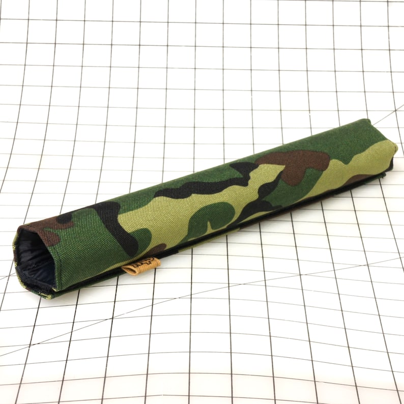 Puede incluir: Un tubo de tela con estampado de camuflaje, una cremallera negra y una etiqueta de cuero marr&oacute;n. El tubo est&aacute; sobre un fondo blanco cuadriculado.
