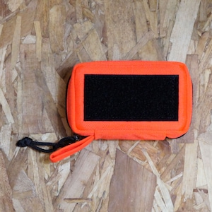 Puede incluir: Una bolsa naranja brillante con cremallera, solapa de velcro negra y correa negra. La bolsa tiene un pequeño parche negro en la parte delantera.