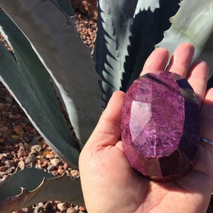 May include: A large, faceted, purple gemstone with a smooth, polished surface. The gemstone is held in a hand, with the background out of focus.