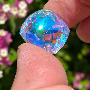 May include: A faceted, square-cut gemstone held between fingers. The gem displays a spectrum of colors, including blues, greens, and pinks, reflecting light. The background features blurred green foliage and pink flowers.