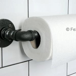 May include: Black industrial pipe paper towel holder mounted on a white tiled wall. The holder has a round flange with the number "1949" on it. The paper towel roll is white.