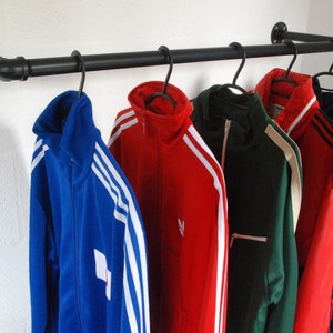 May include: Five track jackets hanging on a black metal pipe rack. The jackets are in various colours, including blue, red, green, and black. Each jacket has white stripes down the sleeves and a white logo on the front.
