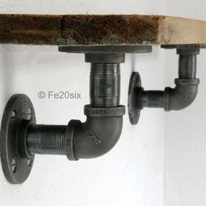 May include: A rustic wooden shelf with two black metal pipe brackets. The brackets are attached to the wall with flanges and have a 90-degree elbow.