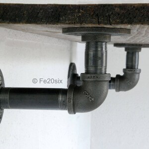May include: A black metal shelf bracket with a wooden shelf. The bracket is made of industrial pipe fittings and has a flange on the wall side.