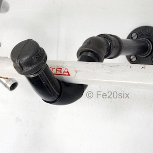 May include: A black metal bike rack with a white painted bar and a black pipe fitting. The rack is mounted to a white wall. The text "Fe20six" is visible on the bar.