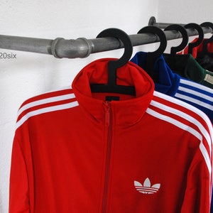 May include: A wall-mounted pipe rack with several tracksuits hanging on it. The tracksuits are in various colours, including red, blue, green, and black. The tracksuits have white stripes on the sleeves and the Adidas logo on the front.