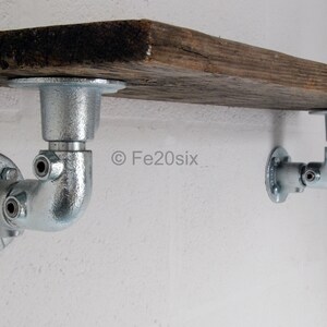 May include: Two silver metal pipe shelf brackets supporting a rustic wooden shelf. The brackets are fixed to a white wall. The shelf is made from reclaimed wood.