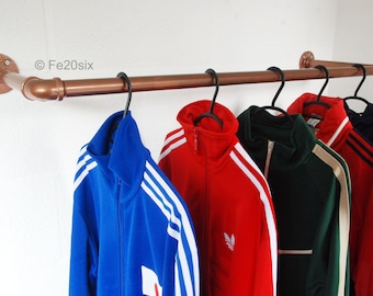 Op de afbeelding: Een kledingrek van koperen pijp met vijf jassen die eraan hangen. De jassen zijn blauw, rood, groen, beige en zwart. Het rek is bevestigd aan een witte muur. ©Fe20six