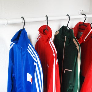 May include: A white pipe clothes rail with black hangers holding several track jackets. The jackets are in various colours, including blue, red, and green. The rail is mounted on a white wall.